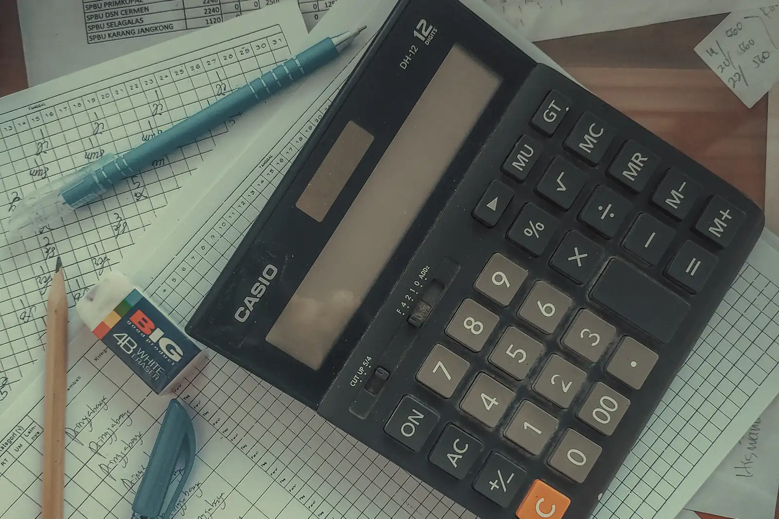 Настольный калькулятор поверх разлинованных таблиц с рукописными записями, рядом карандаш и ручка